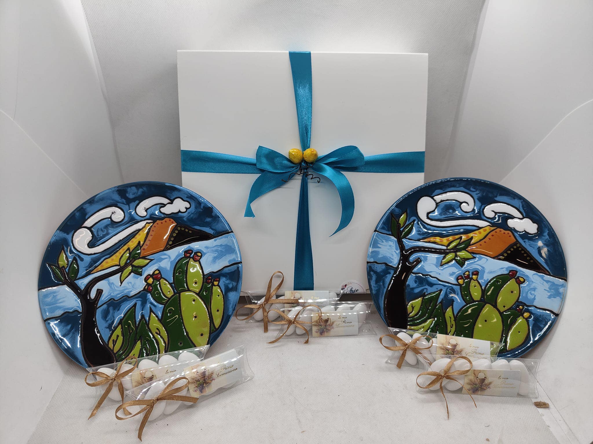 Un assortimento colorato di bomboniere artigianali siciliane in ceramica dipinta a mano. Si distinguono piccole Teste di Moro barute e mini Pigne nei toni del blu pastello, arancio e giallo solare, disposte su un tavolo. Collezioni Bomboniere d'Autore Arte&Decoro a Bronte.