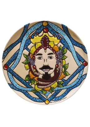 Piatto tondo in ceramica cuerda seca raffigurante una Testa di Moro maschile barbuta incoronata da frutta (limoni, arance, uva) e nastri intrecciati blu. Fatto a mano a Bronte da Arte&Decoro. Elemento scenografico per progetti di interior design.